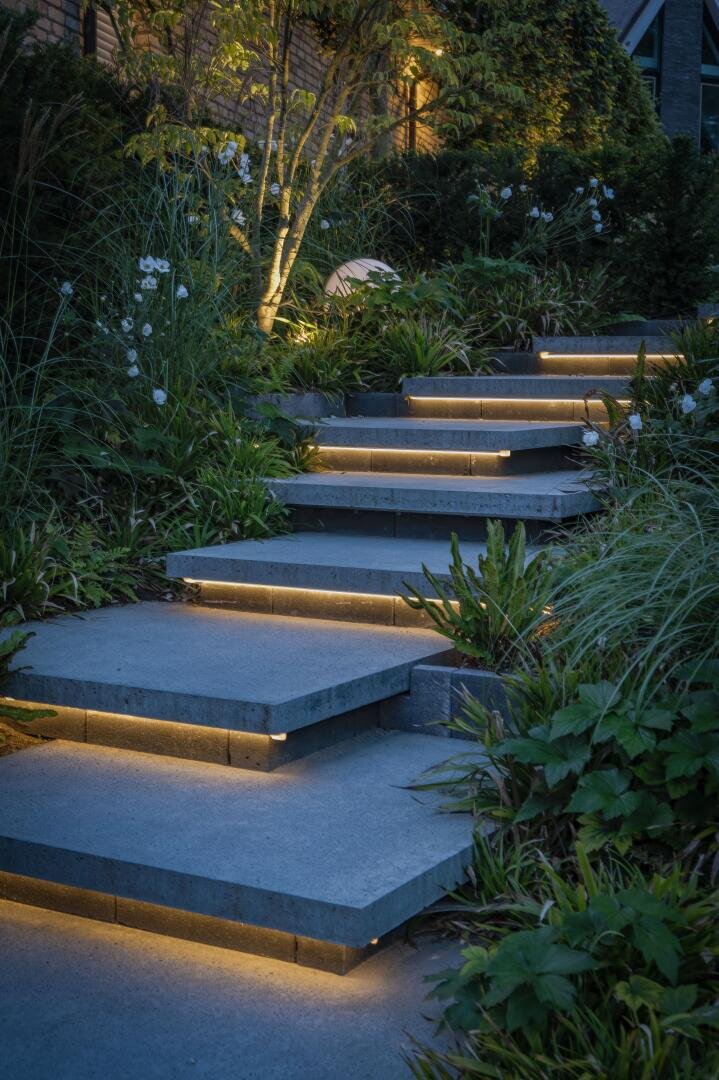 Stone steps with built-in warm yellow lights lead through a lush garden with green plants and white flowers, which becomes an inviting path in the early evening or at night.
