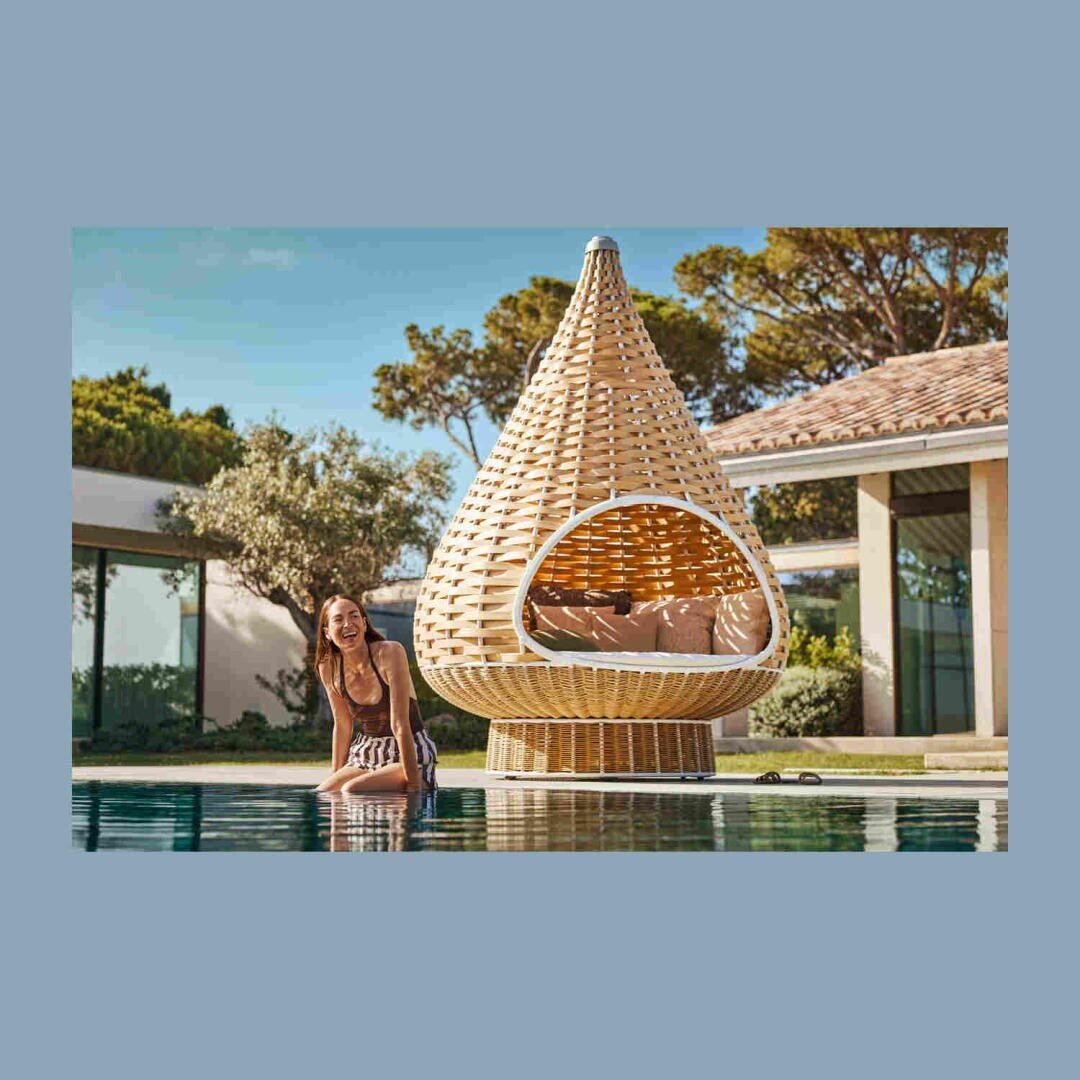 A woman in a swimsuit sits smiling by a pool, behind her a large, teardrop-shaped wicker lounge pod in a sunny setting next to a modern house.