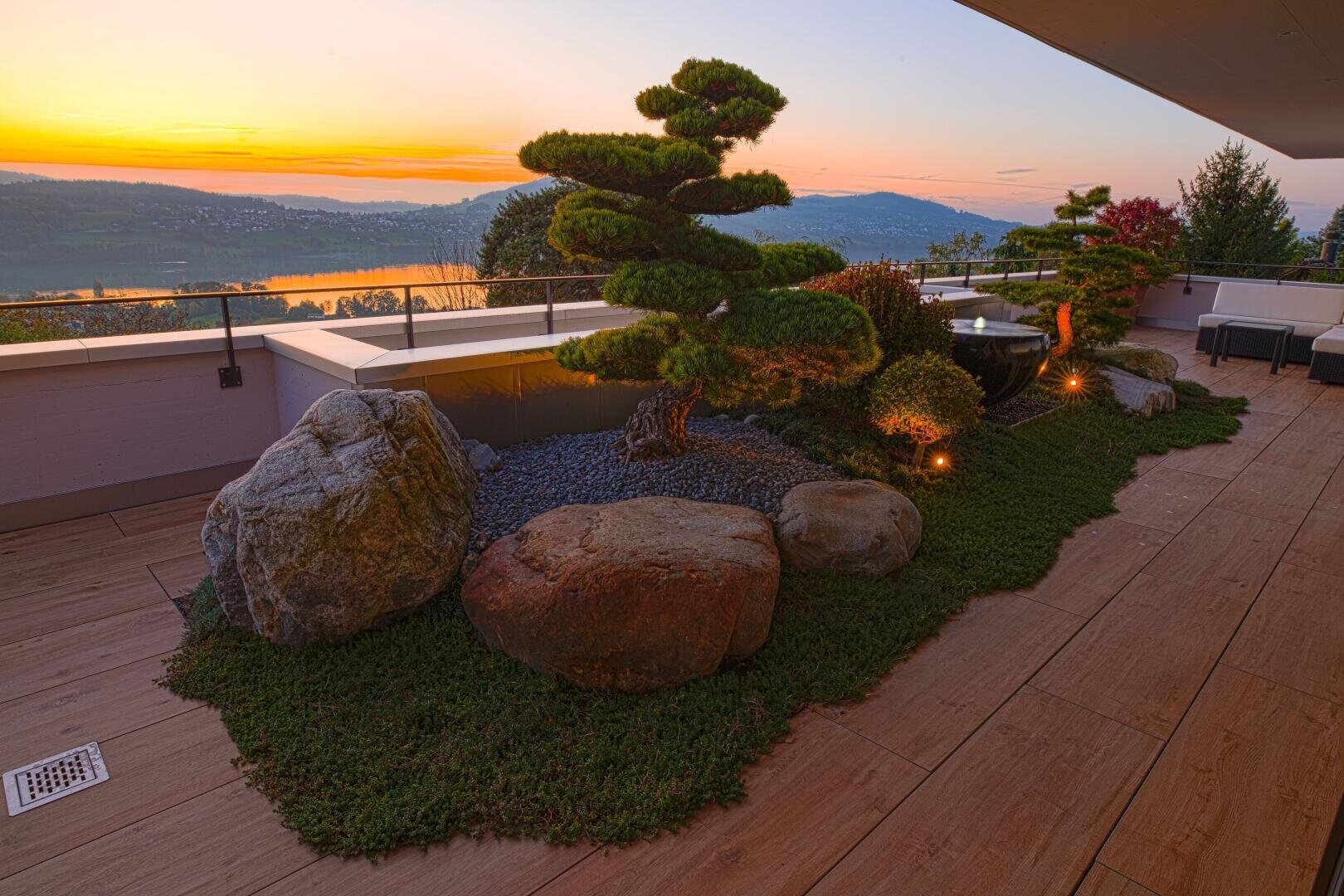 A landscaped terrace with manicured shrubs, large stones and atmospheric lighting offers a view of a picturesque lake and the hills at sunset with warm orange and purple hues in the sky.