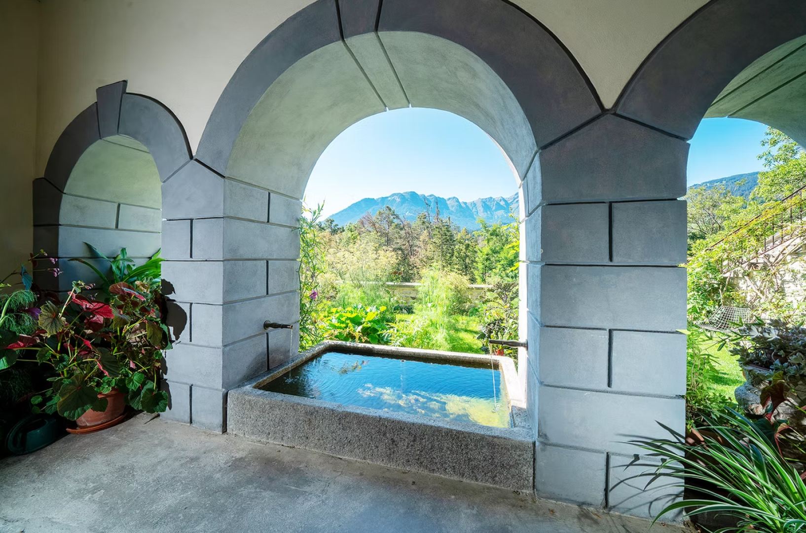 A stone archway frames a small rectangular pool with clear water, surrounded by lush green plants and offering a view of trees and distant mountains under a bright blue sky.