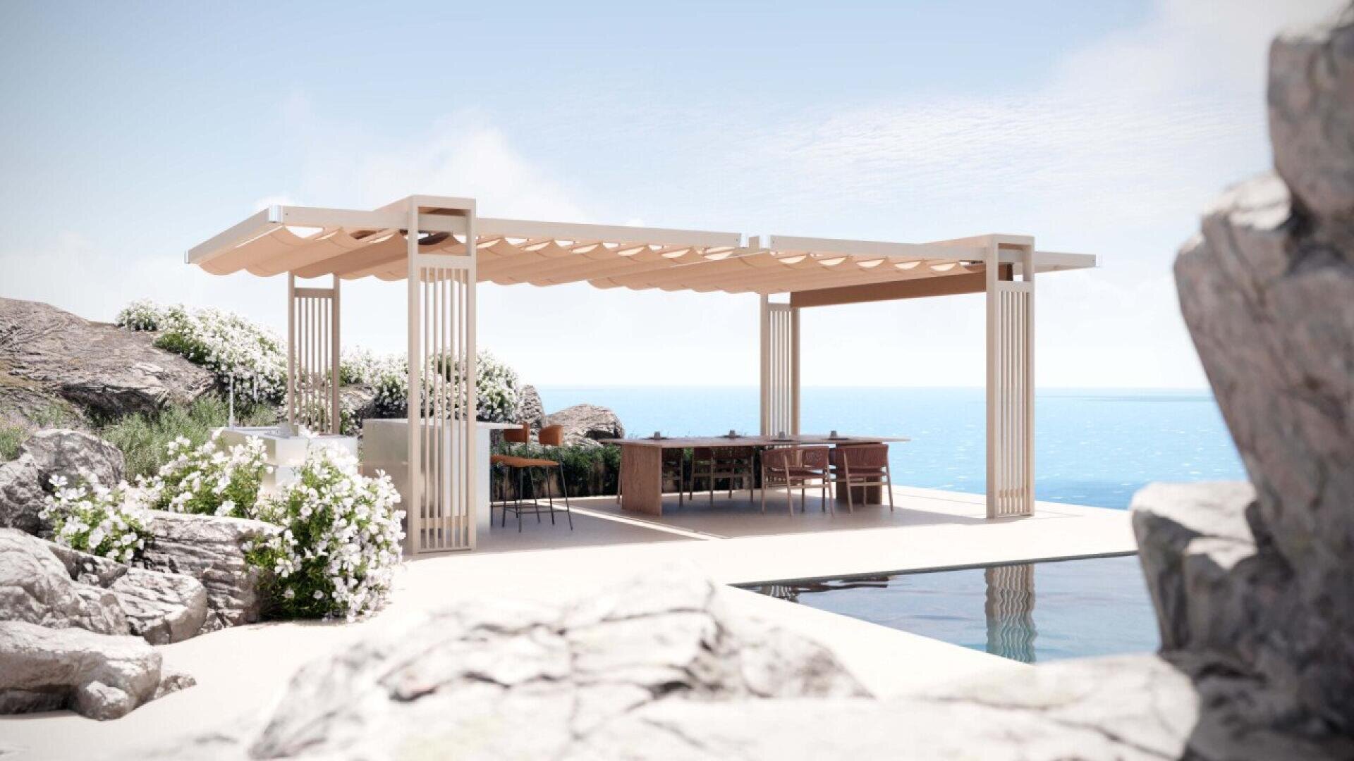 A modern open-air pergola with a long dining table and chairs overlooks the ocean, surrounded by rocks and flowering bushes under a clear blue sky.