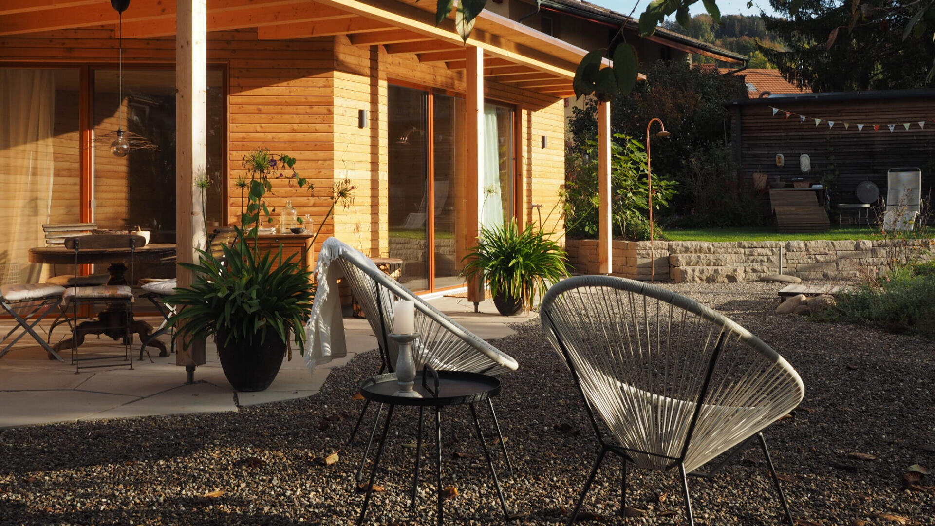 Eine sonnige Terrasse mit modernen Korbstühlen und einem kleinen Tisch befindet sich vor einem Holzhaus. Topfpflanzen schmücken den Bereich, und im Hintergrund befindet sich ein Garten mit Steintreppe.
