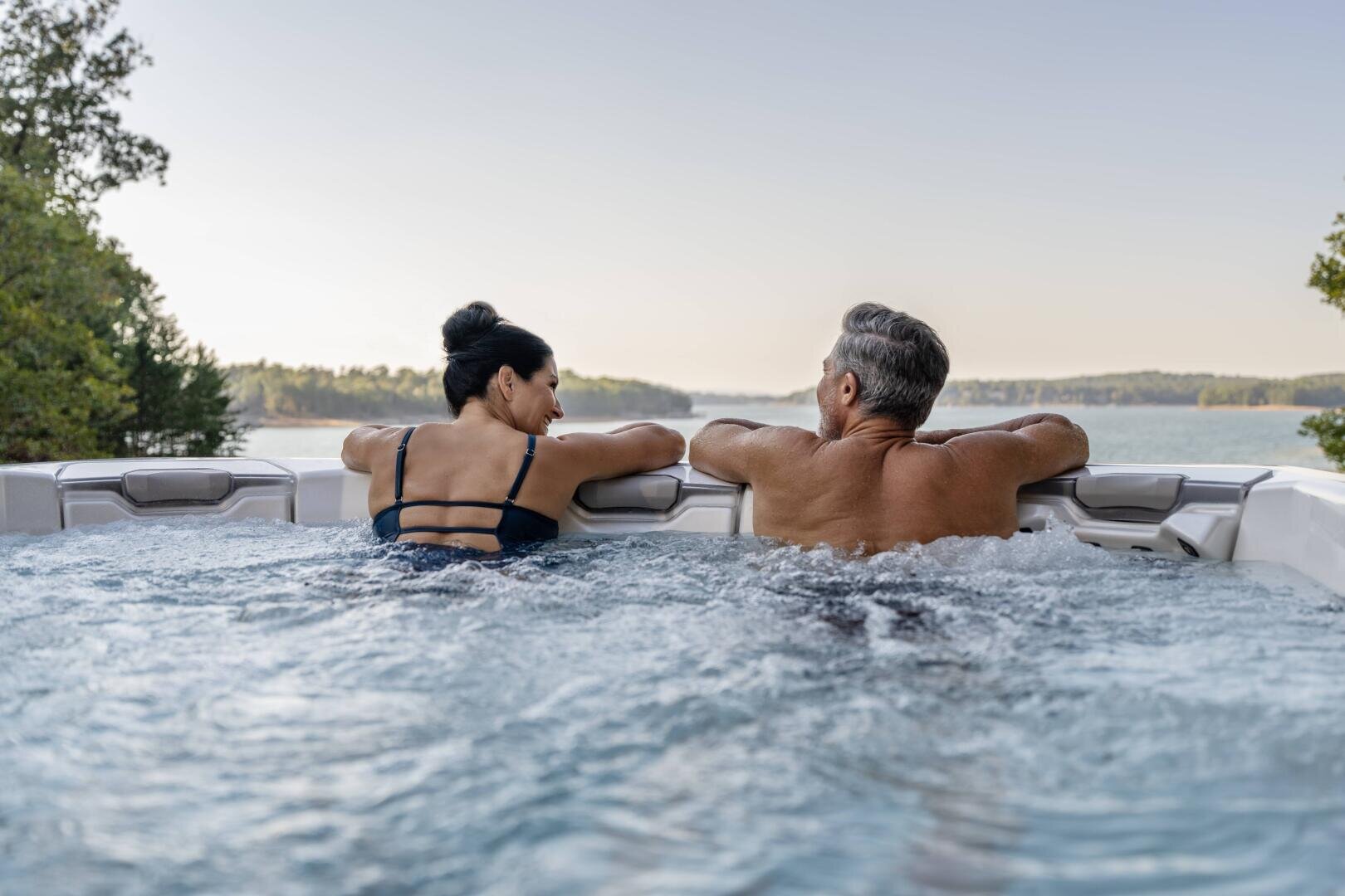 A woman and a man relax in an outdoor hot tub, looking at each other and leaning against the edge. The hot tub overlooks a picturesque lake surrounded by trees under a clear sky.