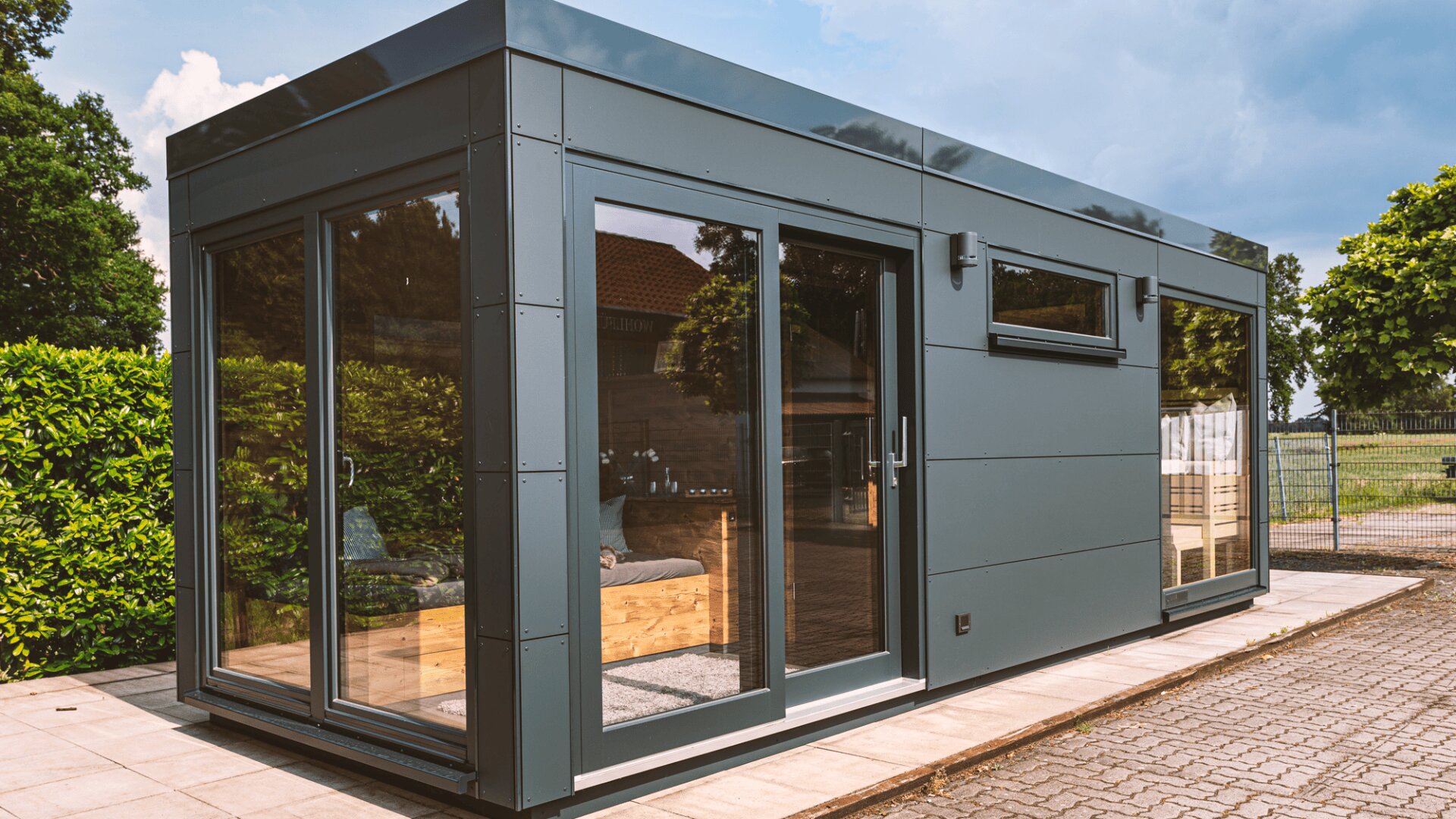 Ein modernes, kleines Haus in Modulbauweise mit großen Glasschiebetüren und -fenstern, einer grauen Außenverkleidung und einem Flachdach, das auf einer gepflasterten Fläche inmitten von Grünflächen und Bäumen steht.
