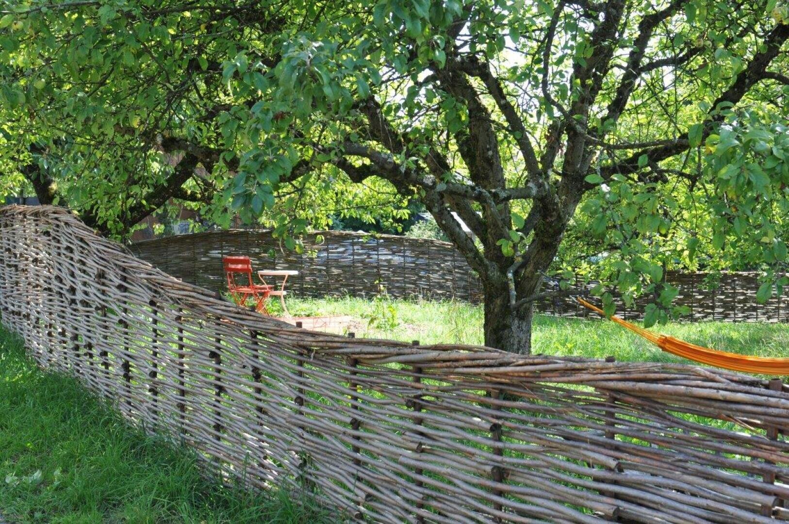Ein rustikaler geflochtener Holzzaun schlängelt sich durch einen grasbewachsenen Garten mit Laubbäumen. Innerhalb des Zauns stehen ein roter Stuhl und ein kleiner Tisch, und auf der rechten Seite ist ein Teil einer orangefarbenen Hängematte zu sehen. Das Sonnenlicht dringt durch das Laub.