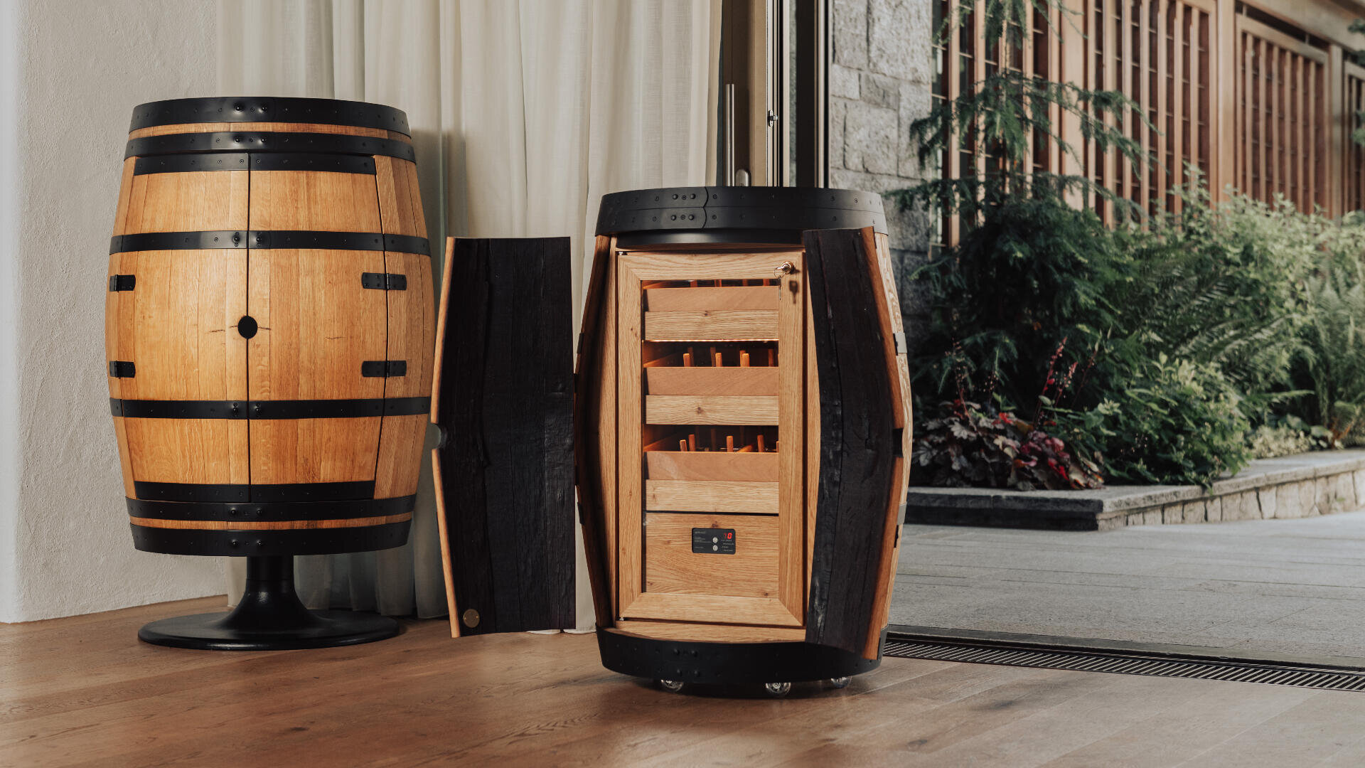 Im Innenraum stehen zwei fassförmige Holzschränke, von denen einer geschlossen und der andere geöffnet ist, um Regale und Stauraum im Inneren freizugeben, ähnlich wie bei einem Wein- oder Schnapsschrank. Eine Schiebetür führt nach draußen in einen Garten.