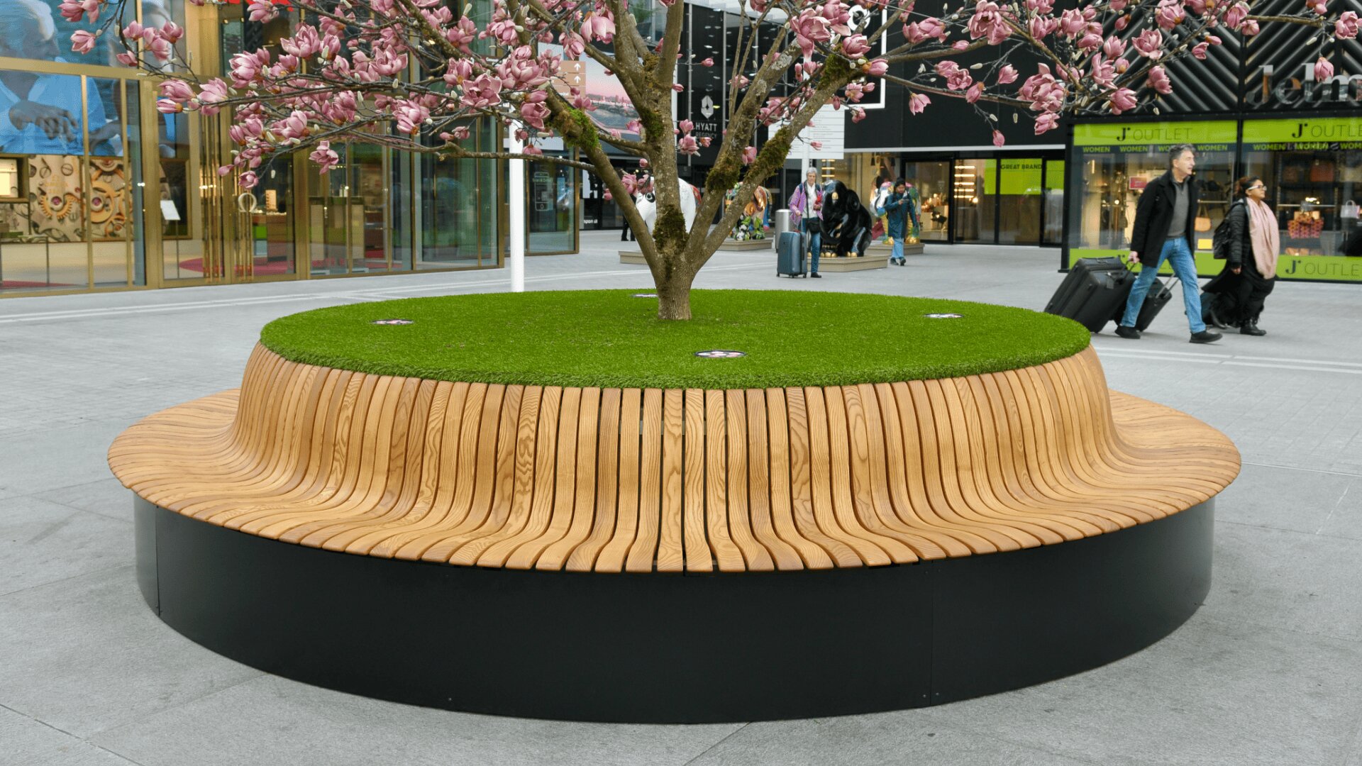 Eine kreisförmige Holzbank umgibt eine Rasenfläche mit einem blühenden rosa Baum in der Mitte, die sich auf einem modernen städtischen Platz befindet, auf dem im Hintergrund Menschen vorbeilaufen.