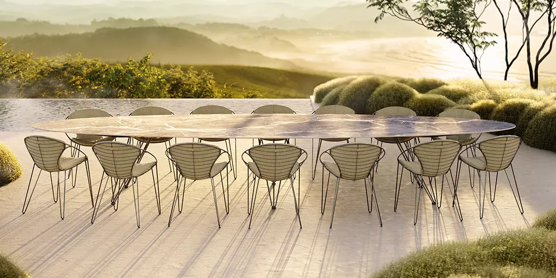 Ein langer ovaler Tisch mit 14 modernen Drahtstühlen steht auf einer steinernen Terrasse, umgeben von grünen Sträuchern, mit Blick auf eine neblige Landschaft mit Hügeln und Bäumen bei Sonnenaufgang oder Sonnenuntergang.