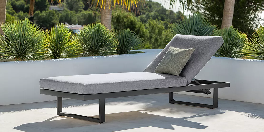 Eine moderne Outdoor-Chaise-Lounge mit grauen Kissen sitzt auf einer Terrasse, umgeben von üppigen Grünpflanzen und Palmen, mit Hügeln und Bäumen im Hintergrund.