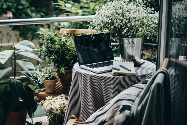 A cozy workspace on the balcony with a laptop, notebook, and pen on a round table covered with a striped cloth, surrounded by lush green plants and white flowers. A chair with a blanket is nearby.