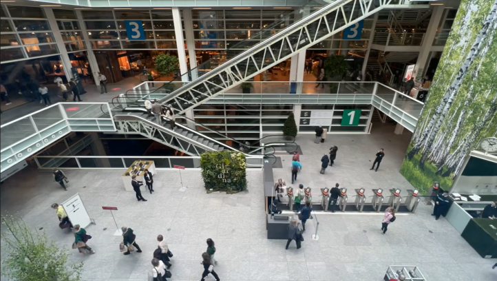 Ein geräumiges, modernes Gebäudeinneres mit Rolltreppen, Glaswänden, nummerierten Schildern und Menschen, die in der Nähe eines Empfangsbereichs gehen oder warten. Große Fenster geben den Blick auf Zimmerpflanzen und die oberen Stockwerke frei.