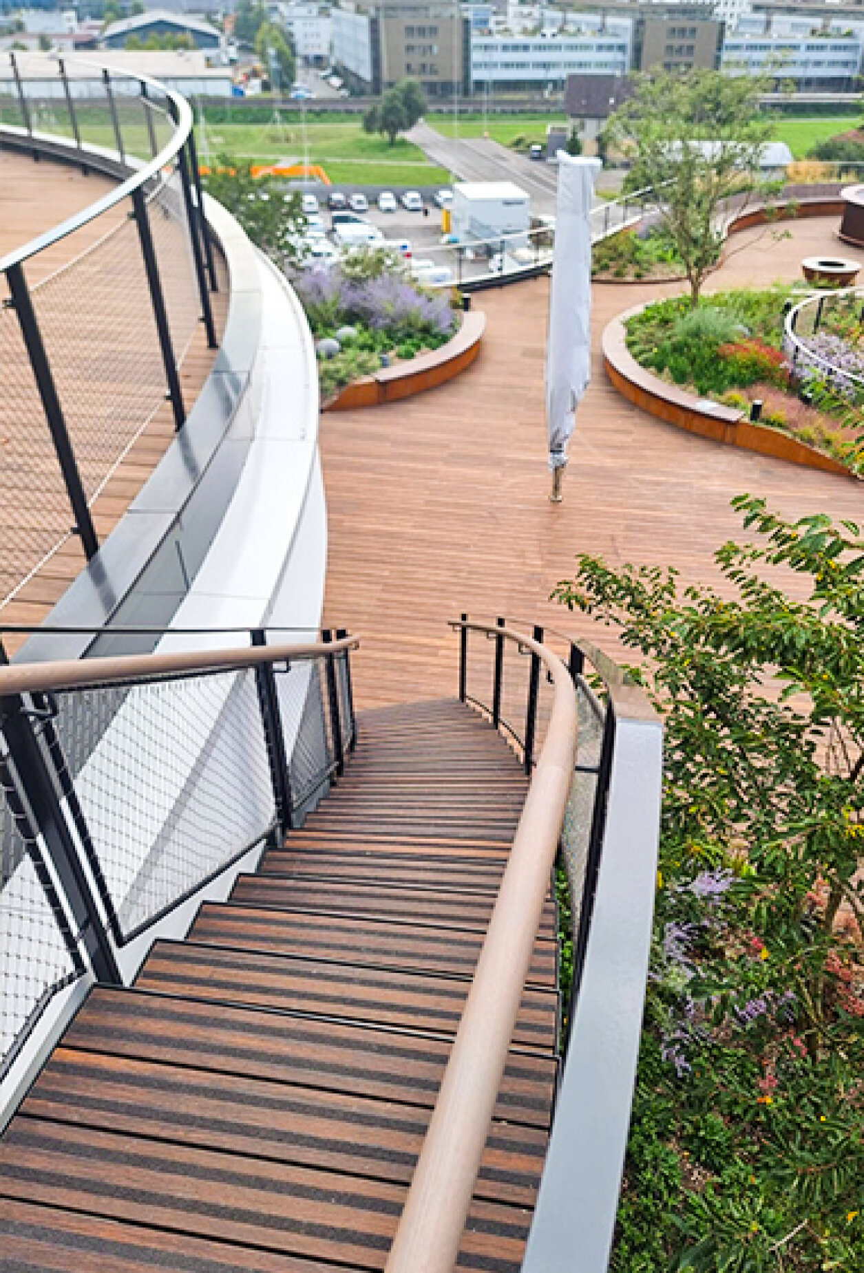 A curved wooden staircase with black railings leads down to a spacious outdoor terrace featuring landscaped gardens and modern seating, with a cityscape visible in the background.
