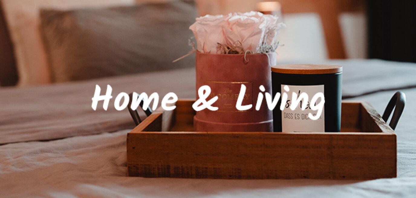Ein Holztablett mit einem rosafarbenen Blumengesteck und einer schwarzen Kerze steht auf einem ordentlich gemachten Bett. Die Worte Home & Living sind in weißer Schrift über das Bild geschrieben.