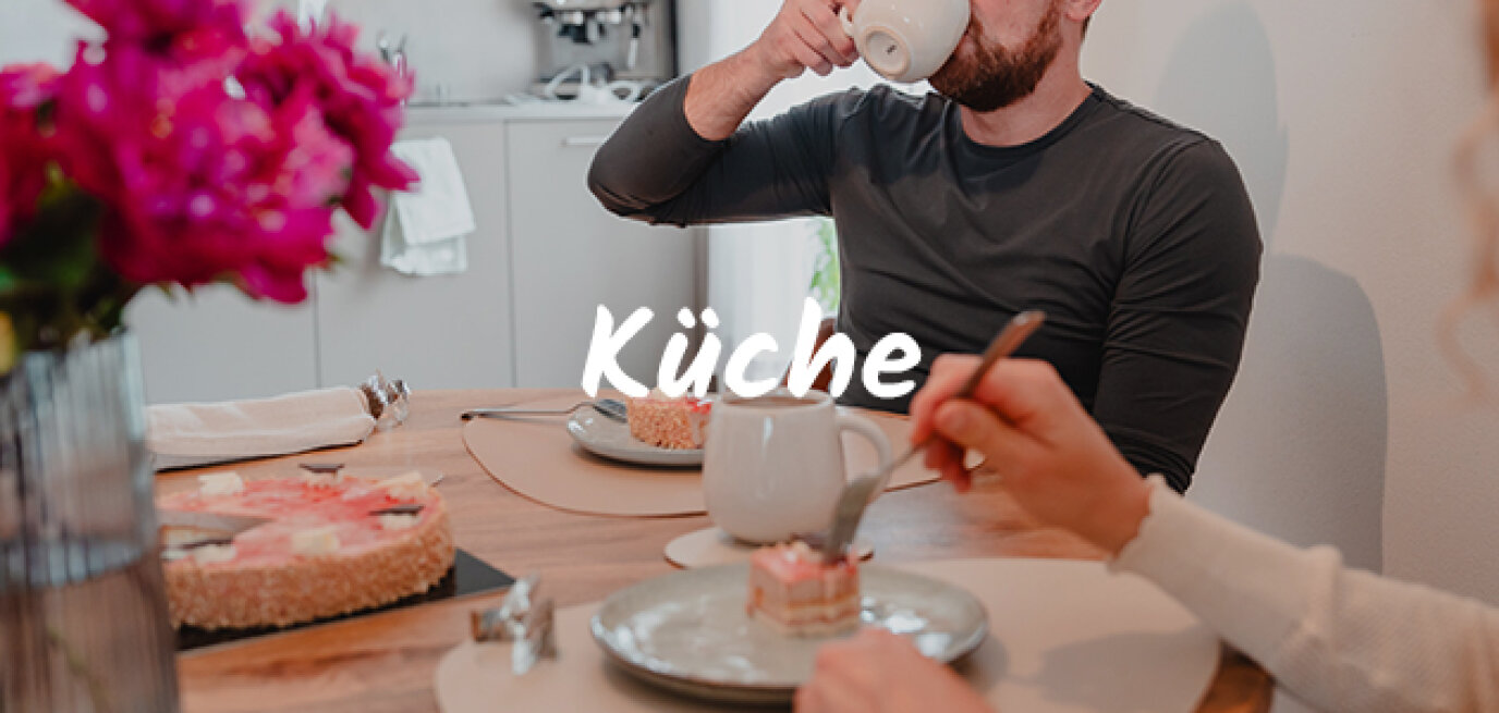 Ein Mann und eine Frau sitzen an einem Küchentisch und genießen Kuchen und heiße Getränke. Im Vordergrund ist ein Strauß rosa Blumen zu sehen. Das deutsche Wort Küche ist quer über das Bild geschrieben.