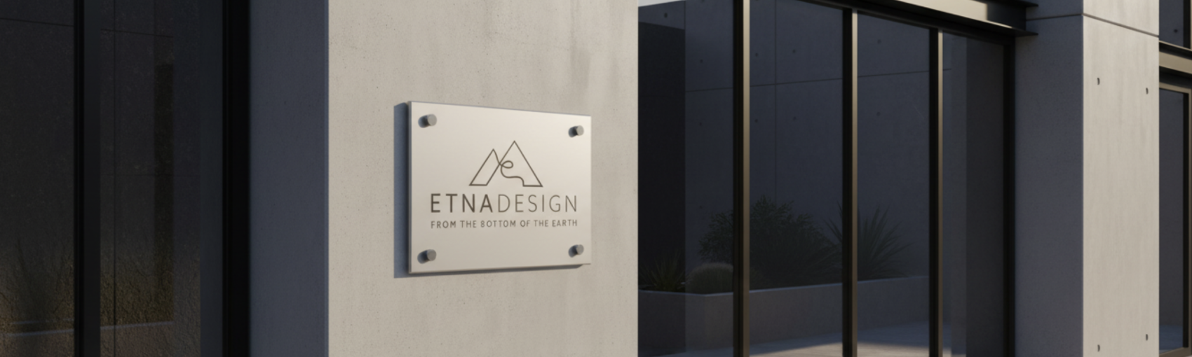 A modern building facade with a metal sign reading ETNADESIGN and the slogan FROM THE BOTTOM OF THE EARTH, along with a minimalist mountain logo, mounted next to large reflective glass windows.