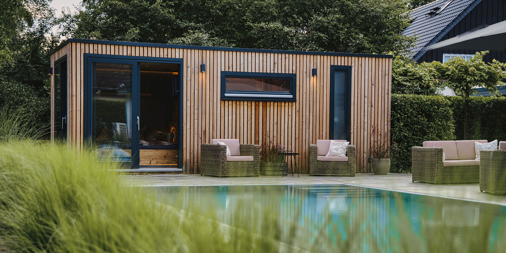 Modernes Holzhäuschen am Pool mit großen Fenstern, umgeben von Korbsofas und Stühlen mit Kissen, üppigen grünen Büschen und einem klaren Swimmingpool im Vordergrund.