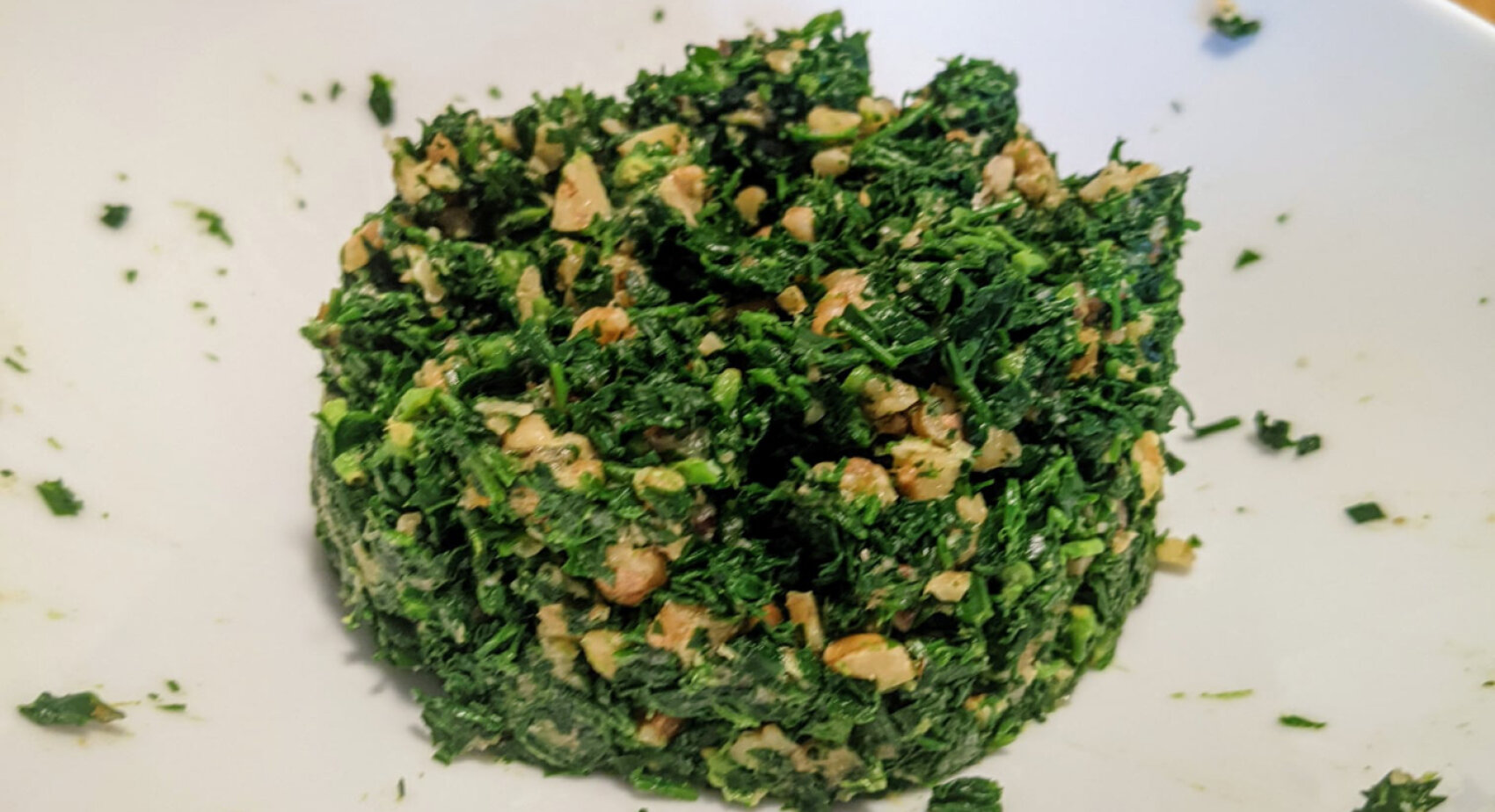 On a white plate is a pile of finely chopped, mixed greens and nuts. The mixture looks fresh and slightly moist, with visible pieces of vegetables and crunchy bits.
