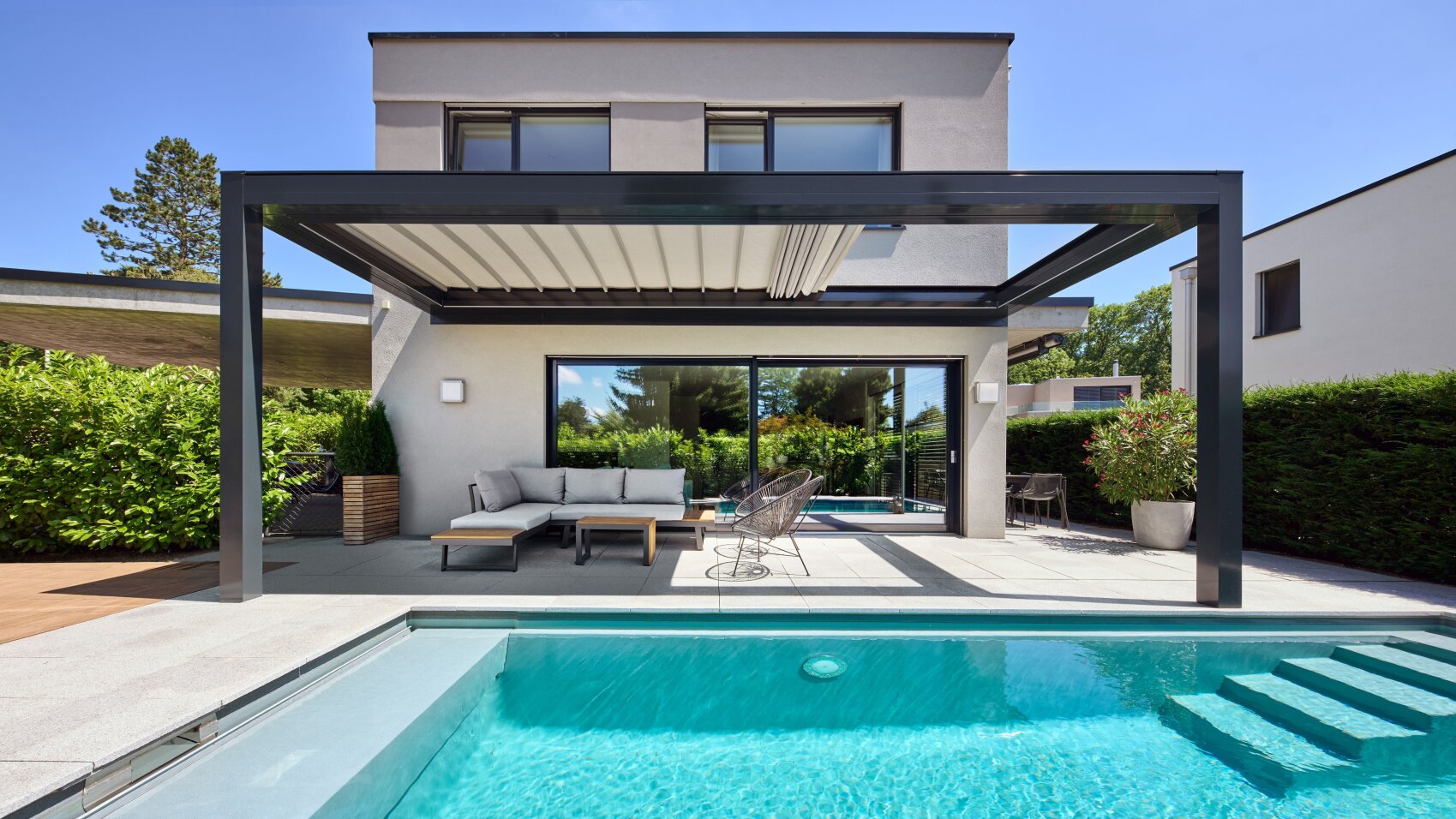 Moderner Garten mit einem Swimmingpool, einer Treppe am Beckenrand, einer schattigen Terrasse mit Sitzgelegenheiten im Freien und einem modernen zweistöckigen Haus mit großen Fenstern im Hintergrund.