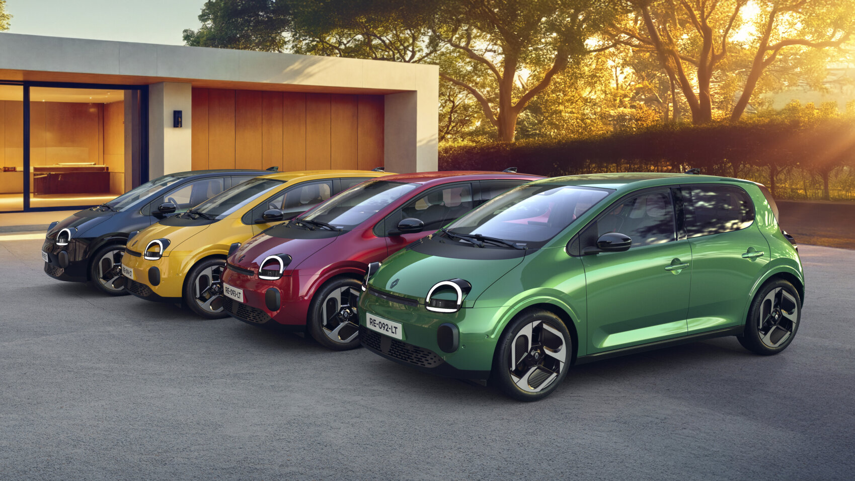 Vier Kleinwagen in verschiedenen Farben - schwarz, gelb, rot und grün - stehen in einer Reihe auf einer Auffahrt vor einem modernen Haus, mit Bäumen und Sonnenlicht im Hintergrund.