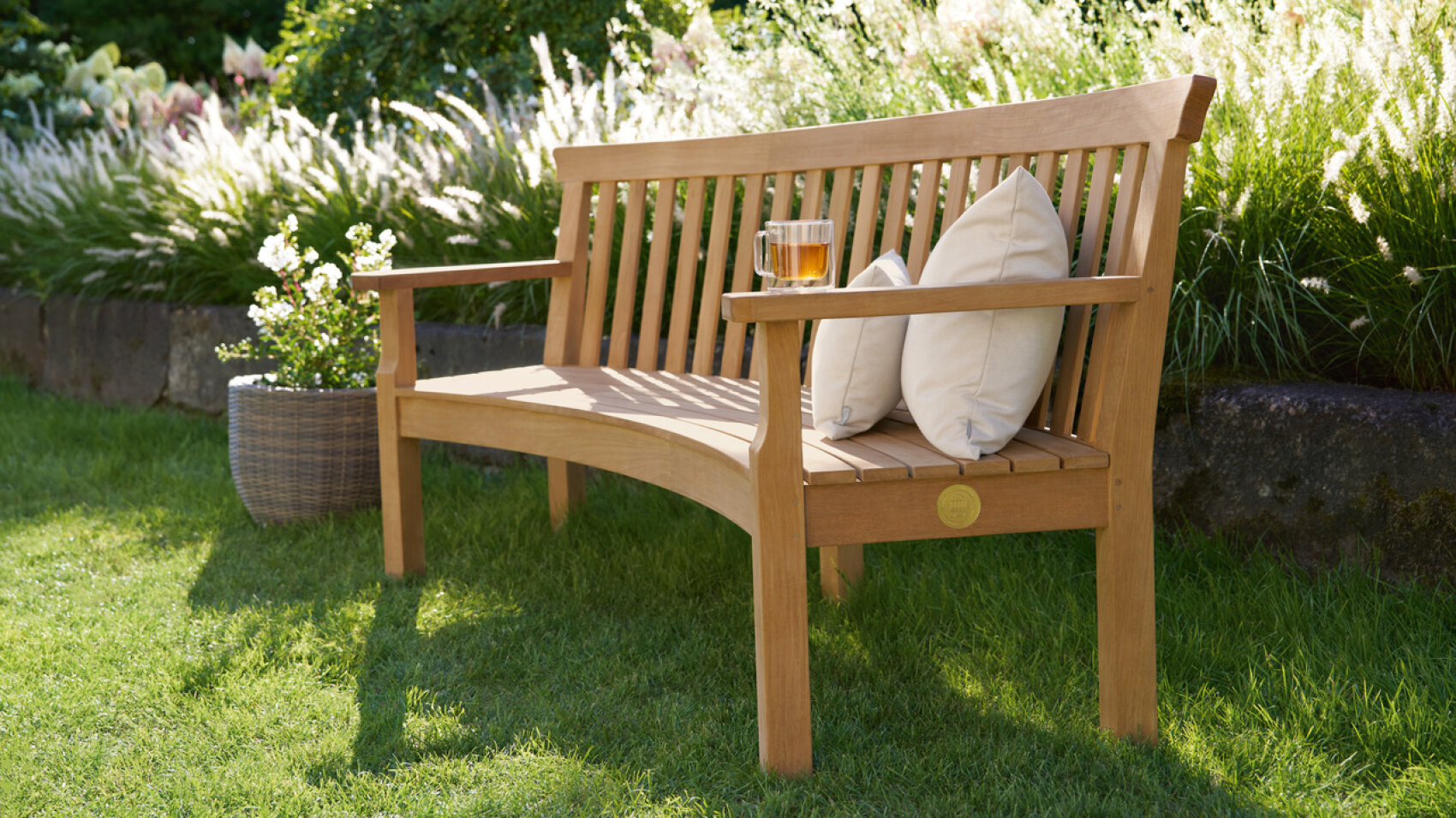 A wooden bench with two white pillows and a glass of iced tea sits on green grass in a sunny garden, next to a basket planter filled with white flowers. Lush plants and flowers are visible in the background.