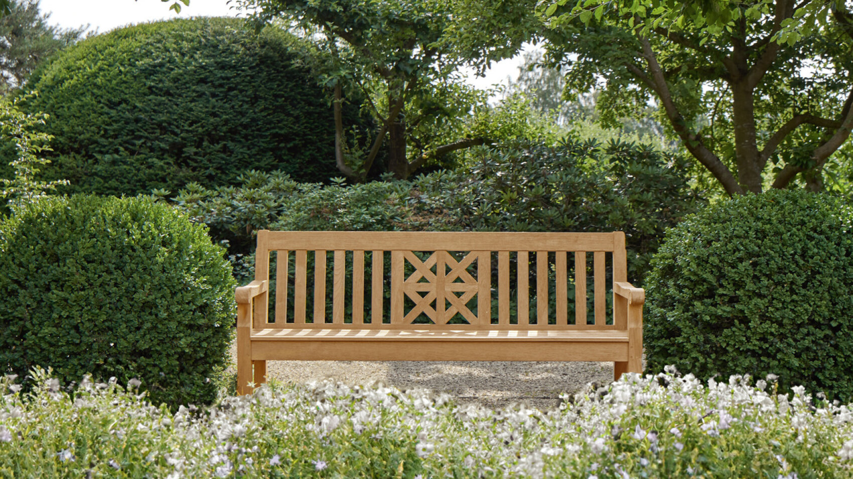A wooden bench with a geometric design on the backrest sits in a garden, surrounded by trimmed bushes, green trees, and flowering plants. The scene is peaceful and inviting.