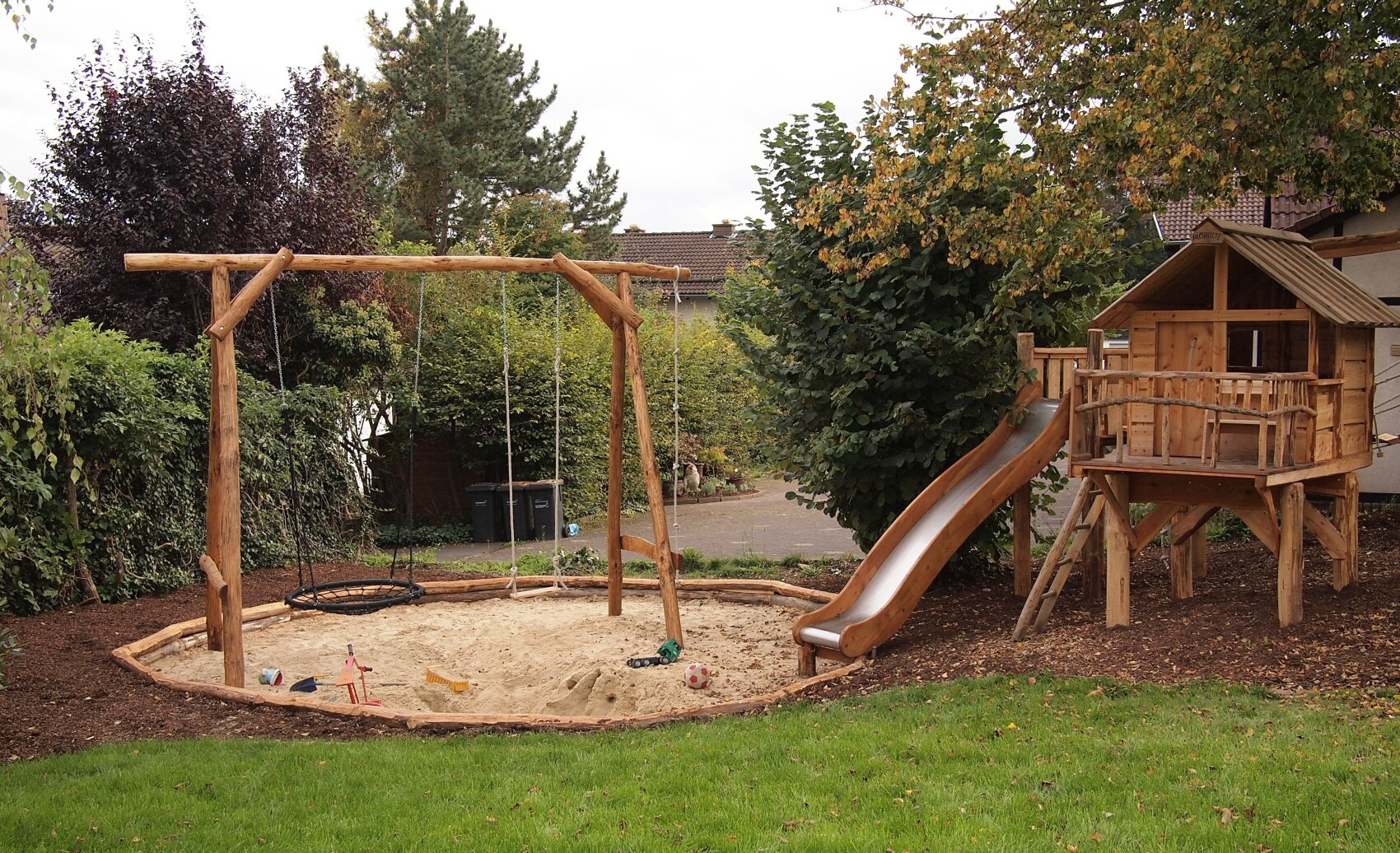 Ein Spielplatz im Hinterhof mit einer Holzschaukel, einem Sandkasten, einer Rutsche und einem Holzspielhaus, umgeben von Bäumen und Gras.