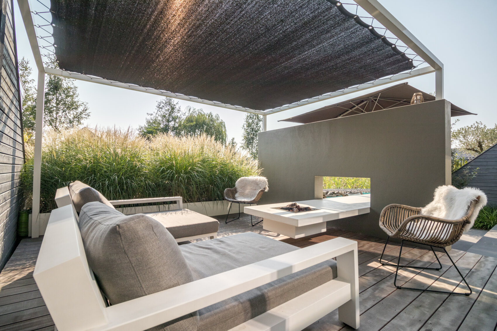 Modern outdoor patio with white sofas, a lounge chair, and a coffee table, featuring a partial wall with an open fireplace. Tall grasses line the back, and a dark shade canopy covers the seating area.