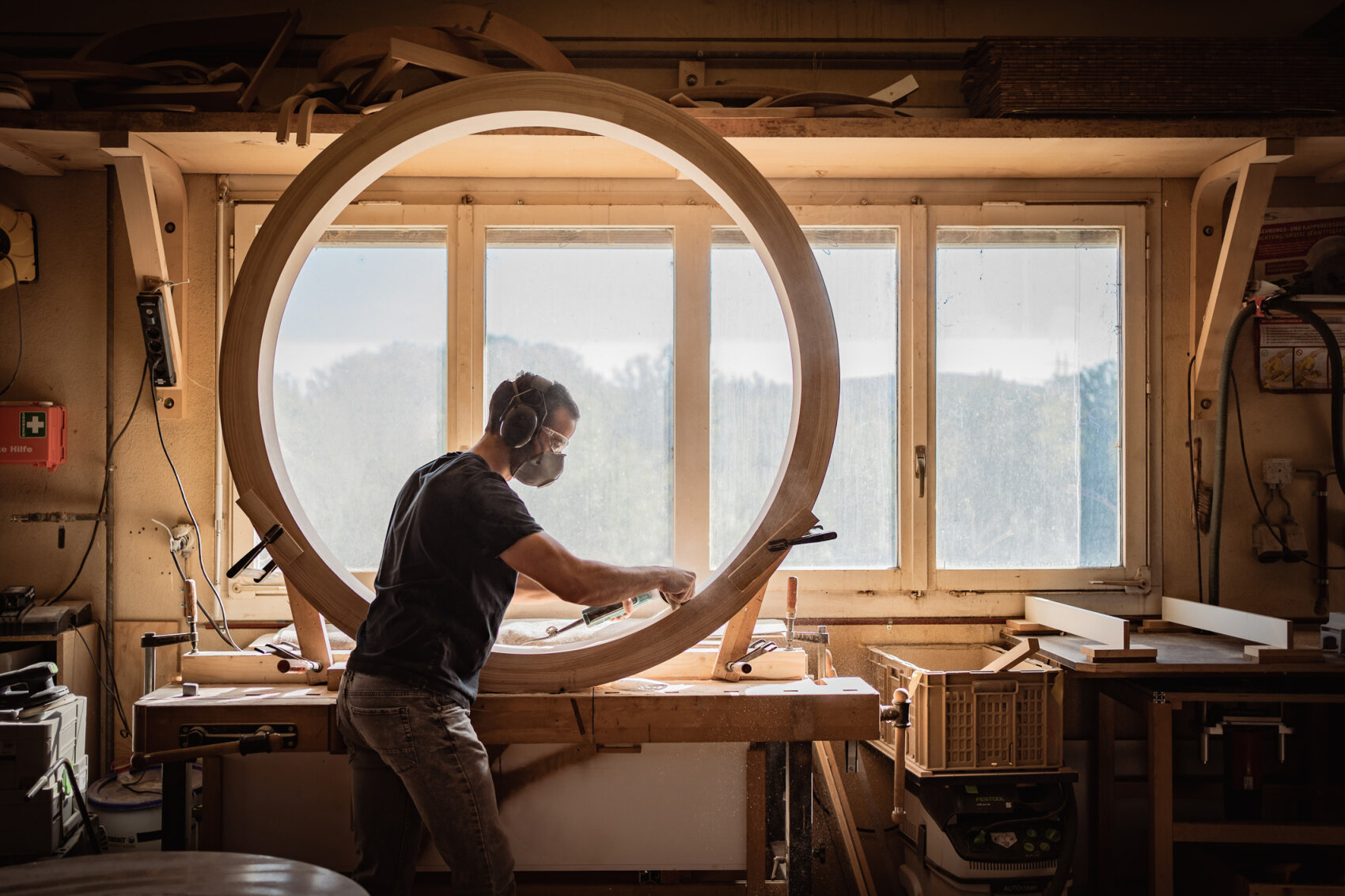 A person wearing headphones and a dust mask works on a large circular wooden frame in a sunlit workshop, with tools and wood pieces on the workbench and shelves.