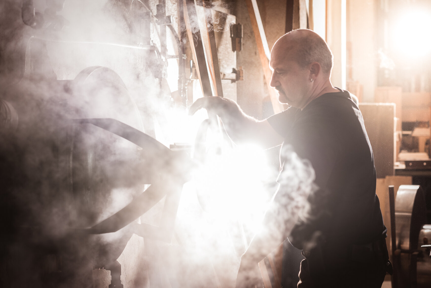 A man operates a large industrial machine surrounded by mist or steam, with bright light shining through, creating a dramatic and atmospheric scene in a factory or workshop setting.