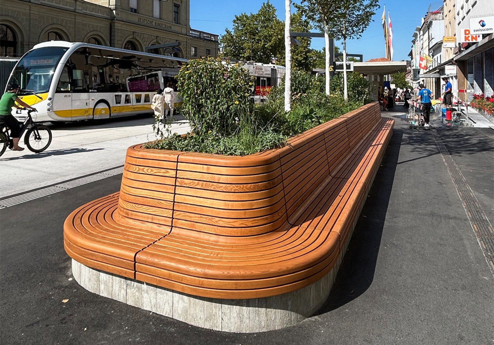 A long, curved wooden bench with built-in planters sits on a city sidewalk, surrounded by pedestrians, cyclists, and passing buses on a sunny day. Buildings and trees line the street.