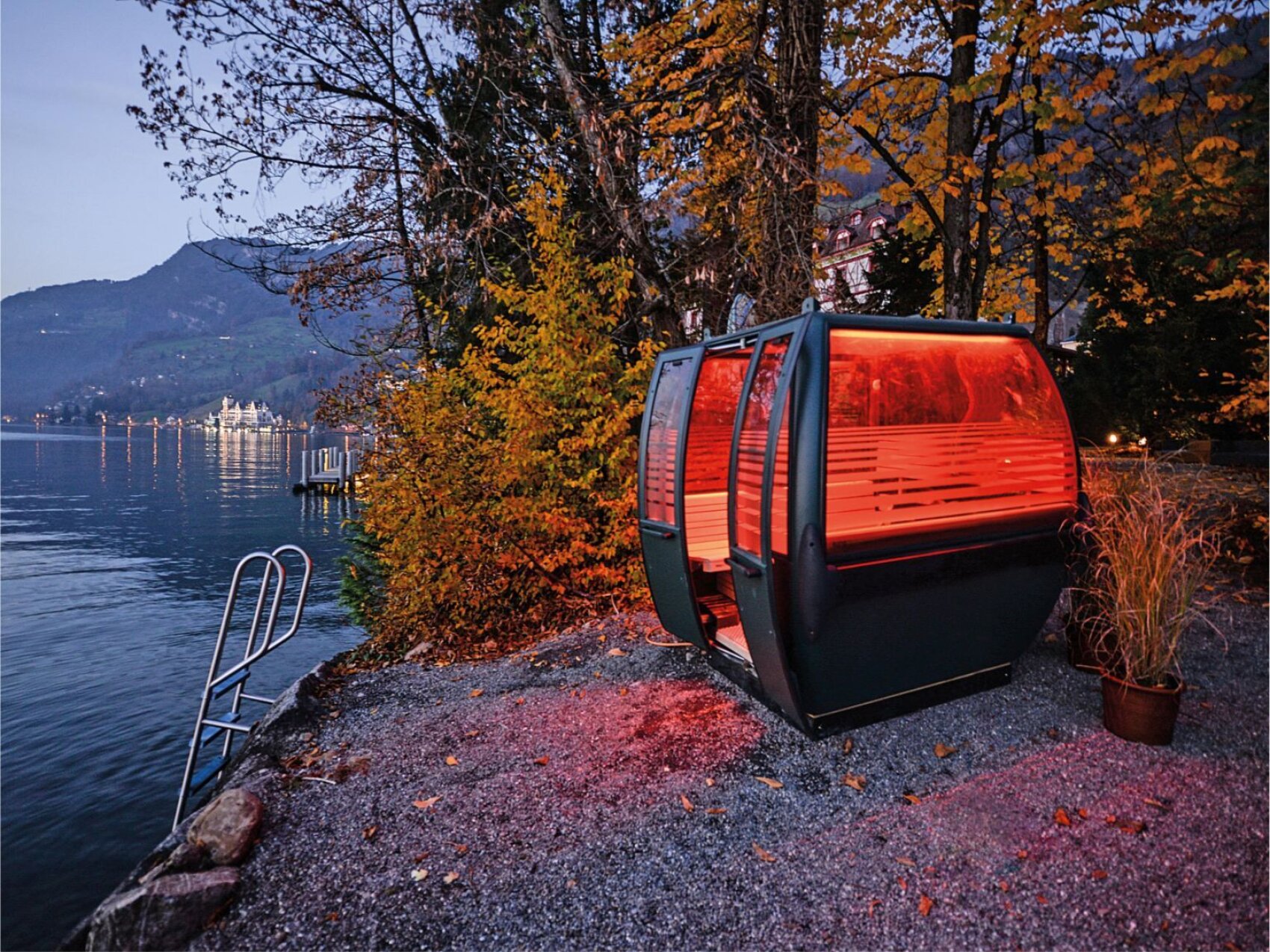 A glowing orange gondola cabin sits by a calm lakeside, surrounded by autumn trees and gravel, with mountains and lights from distant buildings reflected in the water at dusk.