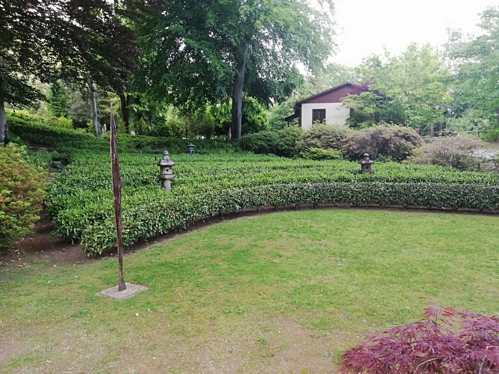 A lush garden with neatly trimmed hedges, green grass, stone lanterns and a small white building in the background, surrounded by tall trees and leafy shrubs.