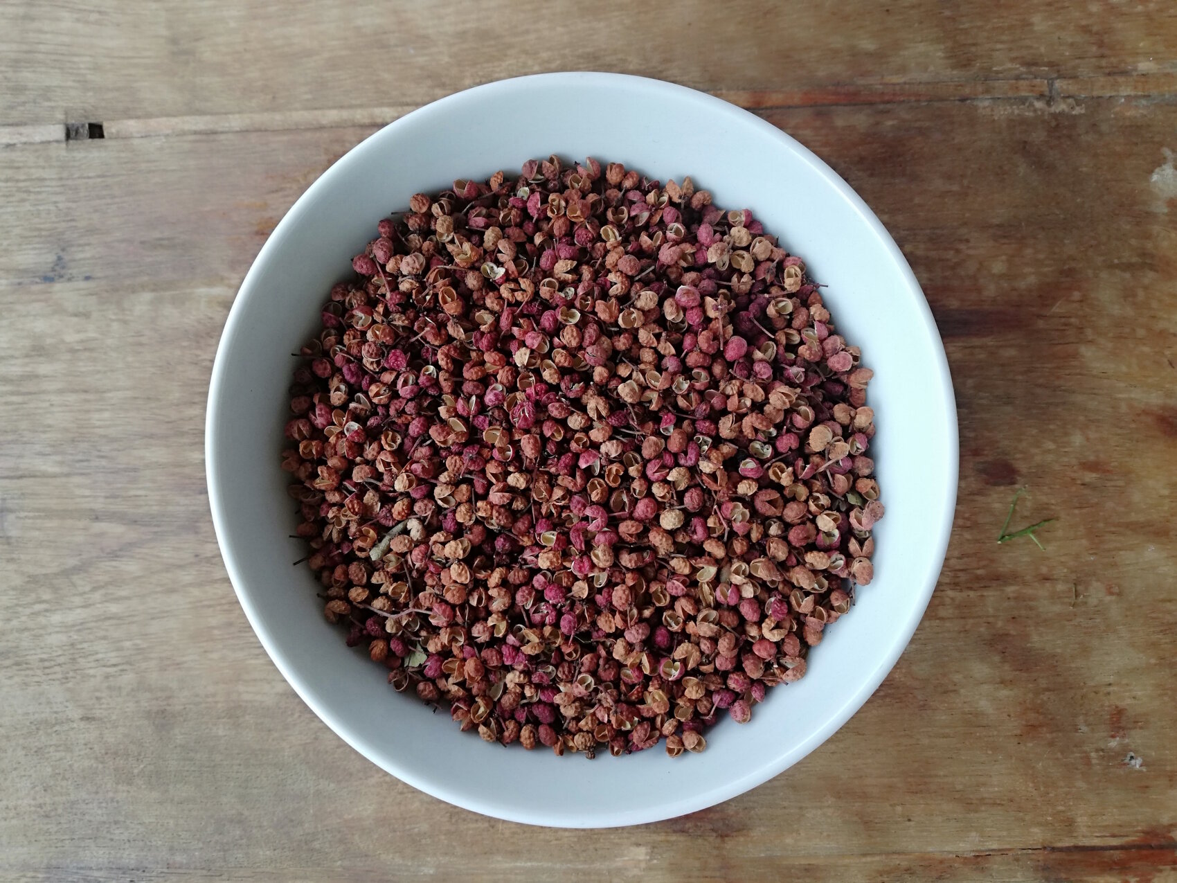 A white bowl of dried Sichuan peppercorns stands on a wooden board. The peppercorns are reddish-brown and round, with a few small stems mixed in.